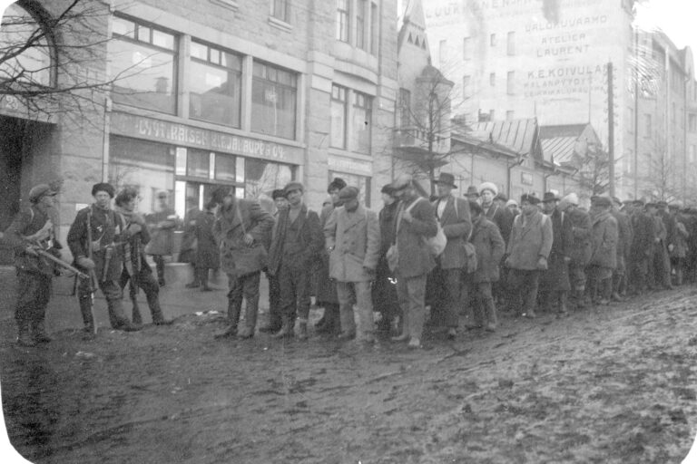 Mustavalkoinen valokuva, jossa miehiä sotilaspuvuissa seisoo joukkona kuvan keskellä jonossa. Ympärillä miehiä aseet käsissä.
