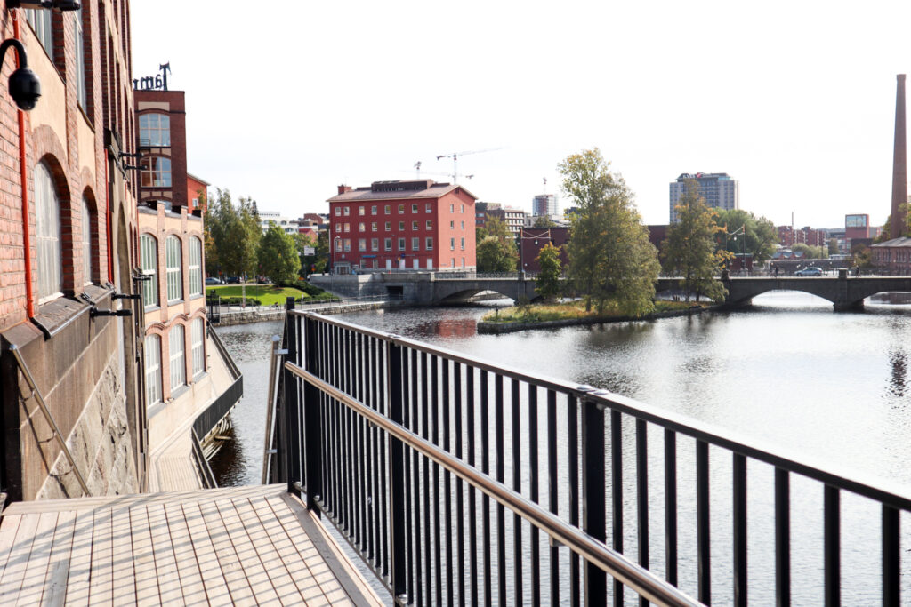 Maisemasilta Vapriikin raitti. Pitkä puinen kävelysilta kulkee Tammerkosken laidalla, toisella puolella punatiilisiä rakennuksia. Kauempana nökyy Tampereen keskustan rakennuksia.