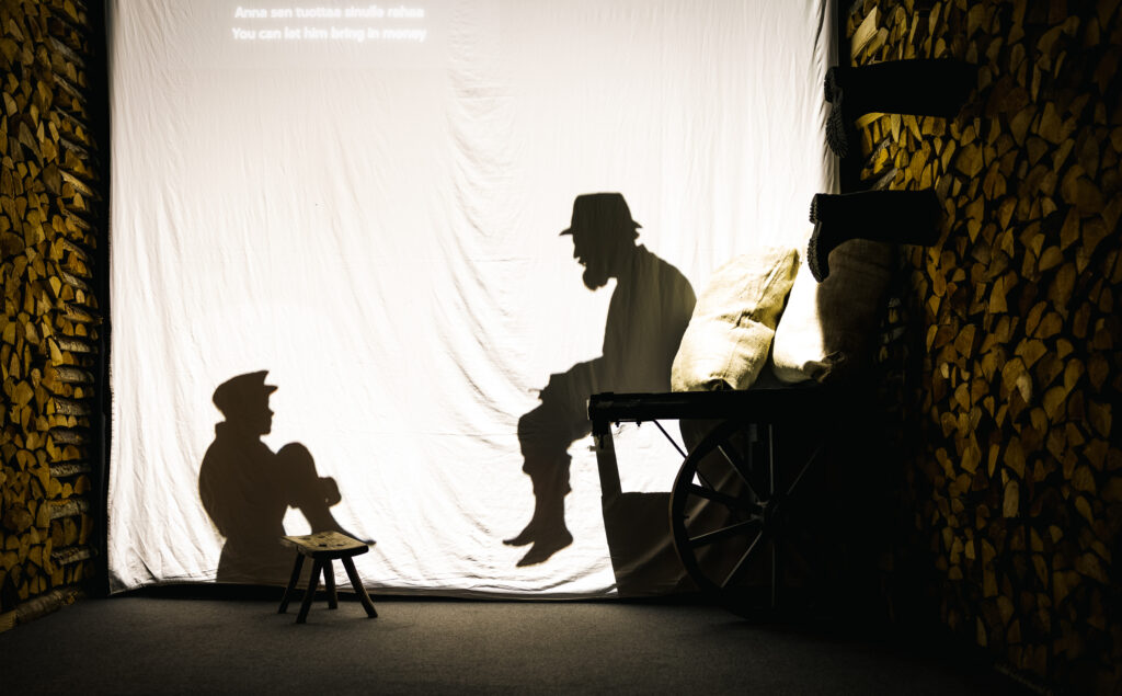 Näyttelytilassa valkoisen kankaan läpi heijastuvat kaksi hahmoa, mies ja pieni poika.