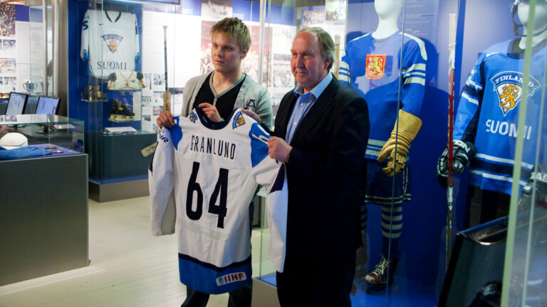 Kaksi miestä seisoo keskellä kuvaa näyttelytilassa. Pitävät käsissä Suomen jääkiekkopelipaitaa jossa lukee Granlund. Takana vitriineissä pelipaitoja.