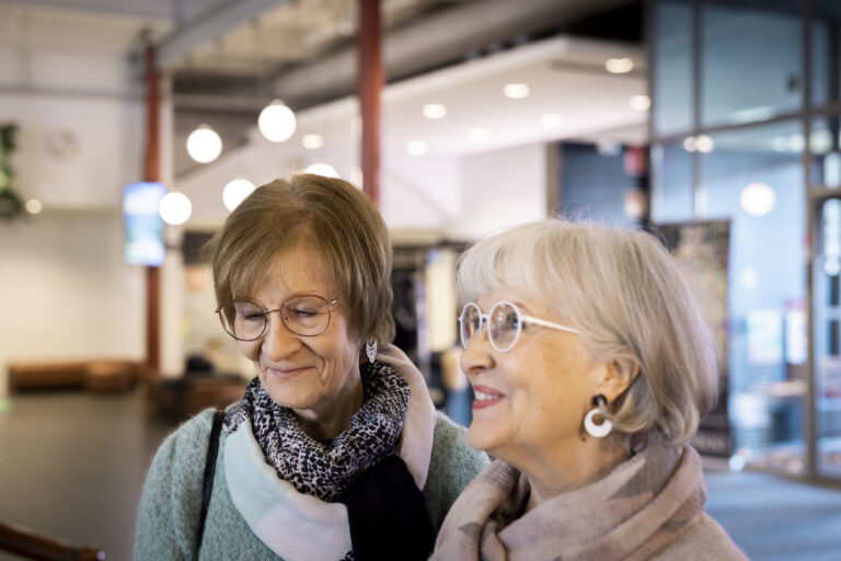 Kaksi senioria Vapriikin aulassa. Toisella naisella on lyhyet harmaan hiukset ja toisella lyhyet ruskeat hiukset. Molemilla on silmälasit.