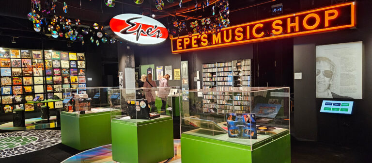 Manserock-näyttelyssä lasivitriinien sisällä esineitä, taustalla levyhylly ja valokyltti, jossa lukee EPES MUSIC.