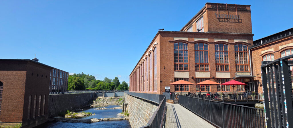 Vapriikin punatiilinen rakennus ulkoa kuvattuna. Edustalla ravintolan ulkoterassi, jossa varjoja ja ruokailijoita. Rakennuksen reunassa alapuolella virtaa koski.