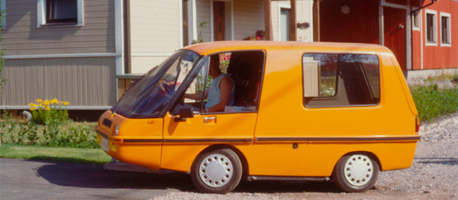 Pieni Postin oranssi sähköauto Finnvan omakotitalon edessä, taustalla vihreä nurmikko ja keltaisia kukkia.