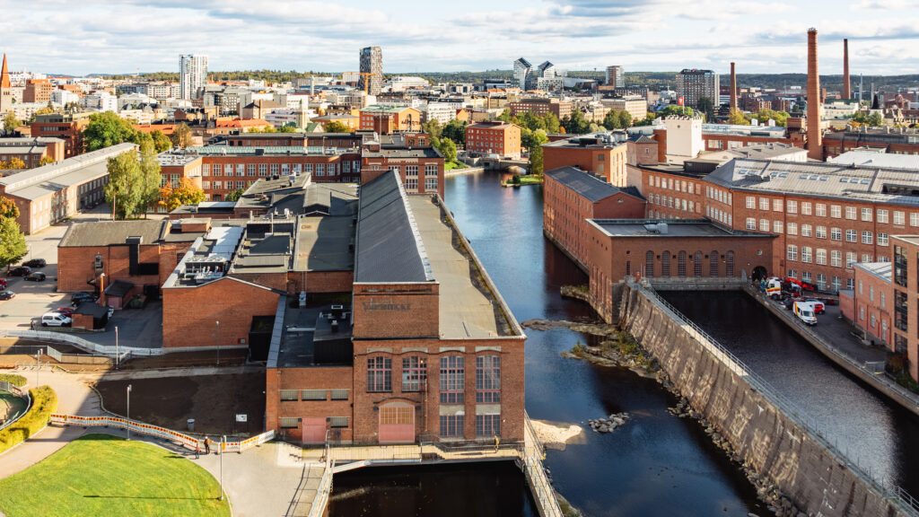 Vapriikin punatiilinen rakennus kuvattuna ilmasta käsin. Rakennuksen toisella reunalla virtaa Tammerkoski. Taustalla Tampereen keskustan rakennuksia,