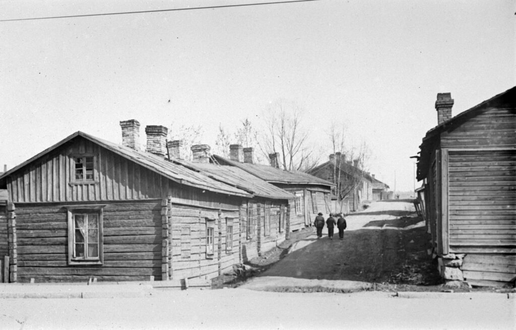 Mustavalkoisessa valokuvassa hirsitaloja kapean hiekkatien molemmin puolein. Tiellä kävelee kolme hahmoa.