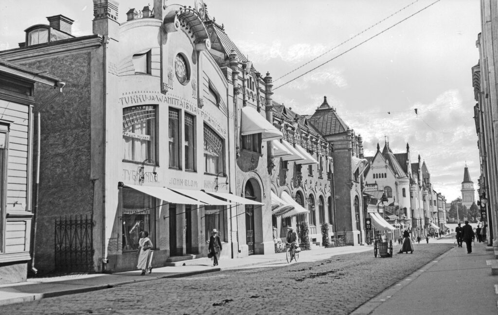 Mustavalkoisessa valokuvassa Tampereen Kauppakadun rakennuksia 1900-luvun alusta. Tiellä kulkee ihmisiä.