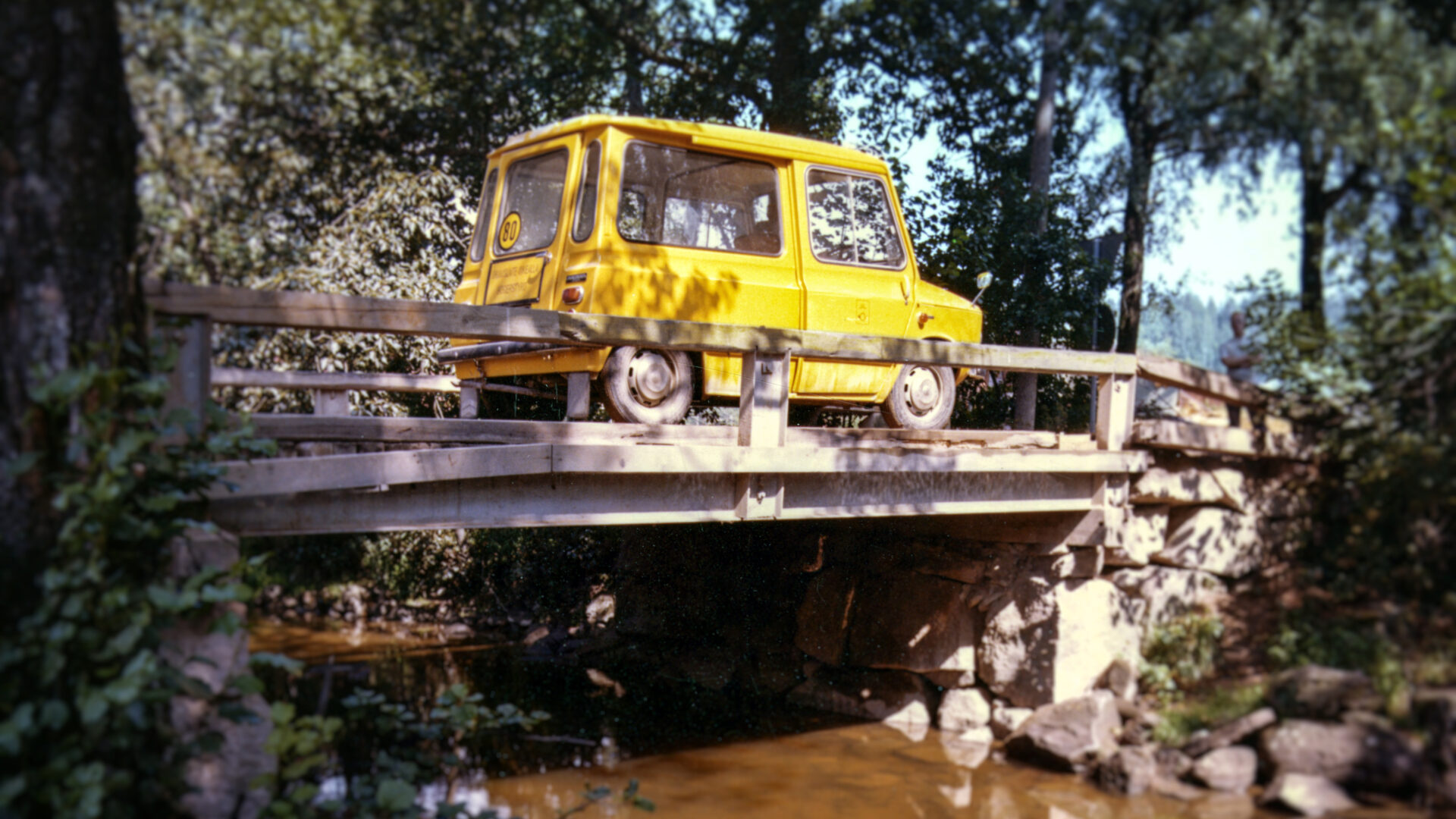 Keltainen postiauti ajaa kapean ja puisen sillan yli metsämaisemassa.