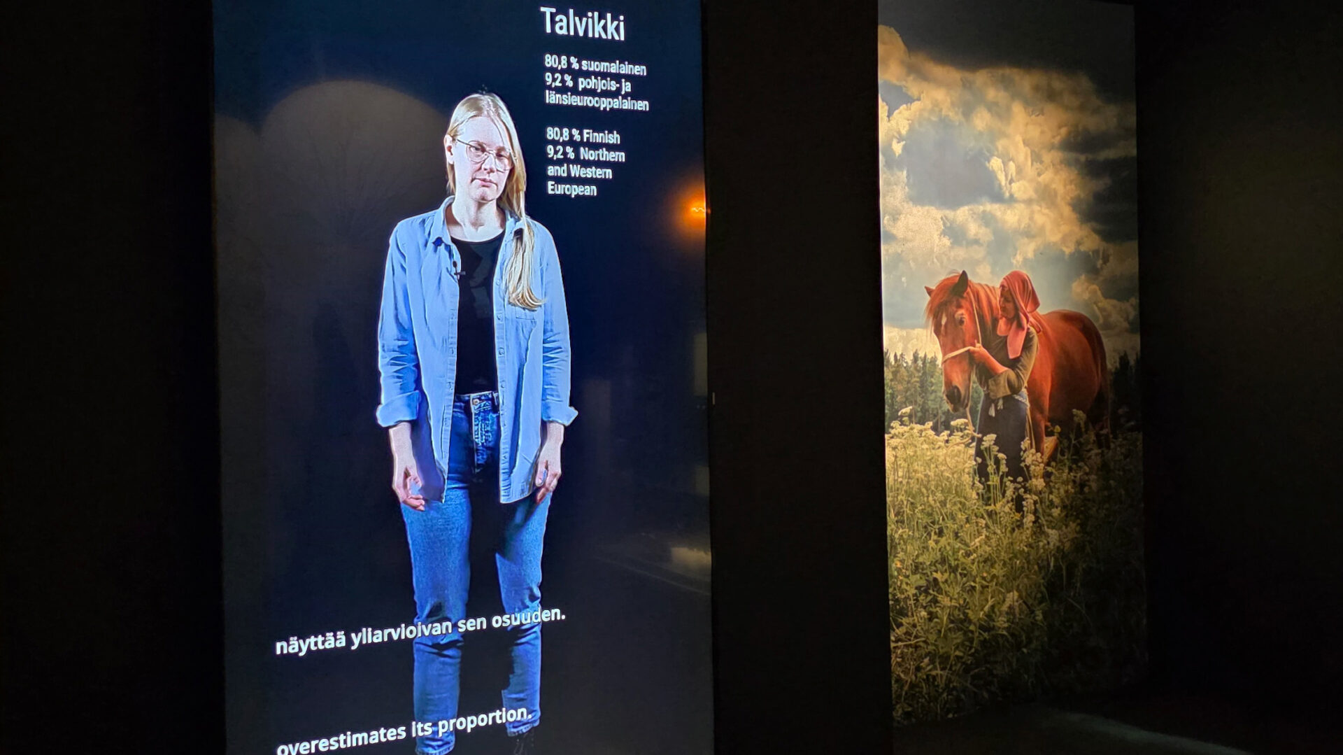 Hämärässä näyttelytilassa näytöllä kuva naisesta. Taustalla seinäkuvassa hevonen.