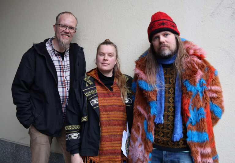 Kolme henkilöä poseeraavat kameralle ulkona, kaikilla takit päällä.