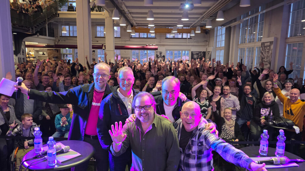 Kuvan edustalla viisi miestä lavalla sisätiloissa vilkuttavat kameralla. Heidä takanaan yleisömeri hurraa.