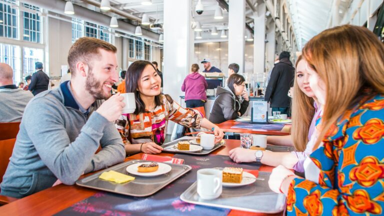 Neljä nuorta aikuista istuu museoravintolan pöydässä vastakkain. Pöydällä leivoksia lautasilla sekä kahvikuppeja.