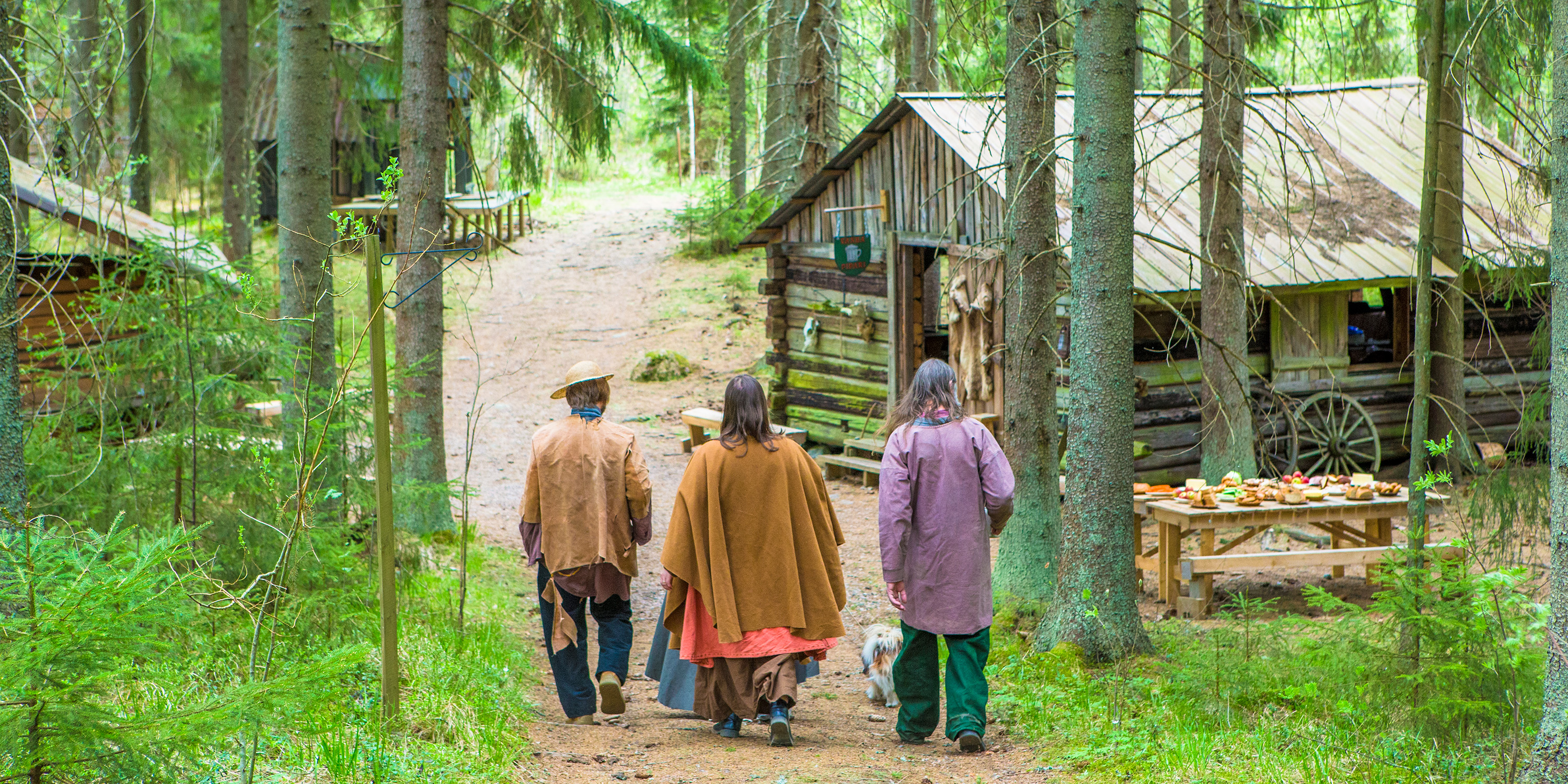 Kesäisessä metsämaisemassa kulkevalla hiekkatiellä kävelee kolme ihmistä kohti edessä olevia puurakennuksia.