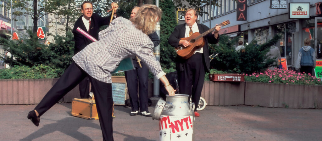 Kolme herraa soittaa ja laulaa taustalla, edessä nainen kumartuneena keräystonkan ylle.