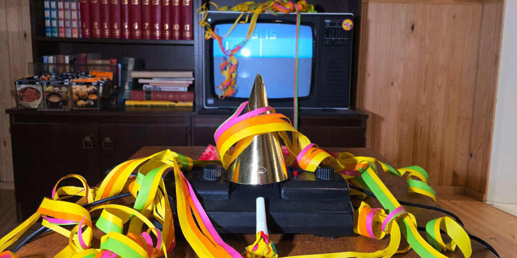 Pelimuseossa ohjain koristeltu serpentiinillä ja juhlahatulla. Taustalla televisooruudulla pyörii peli.