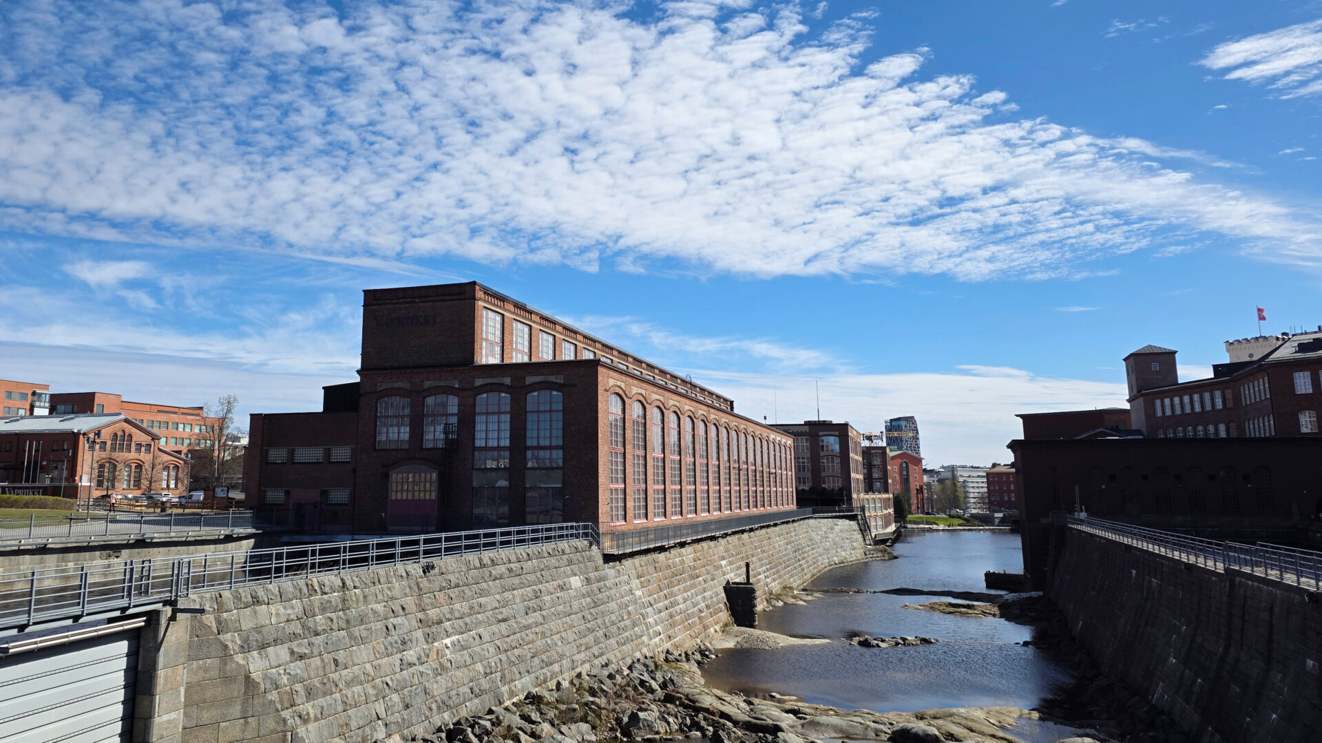 Vapriikin punatiilinen rakennus ulkoa kuvattuna. Rakennuksen reunassa alapuolella virtaa koski.