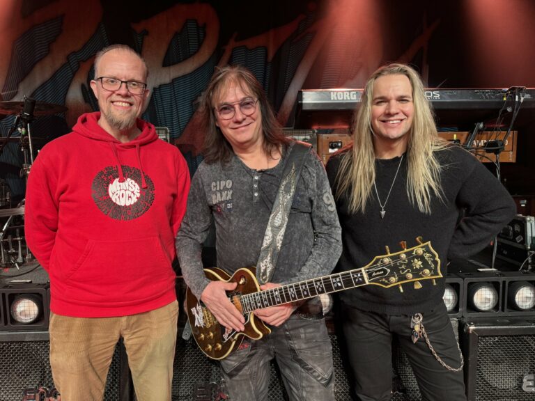 Kolme miestä hymyilee, keskimmäisellä on kädessä kitara.