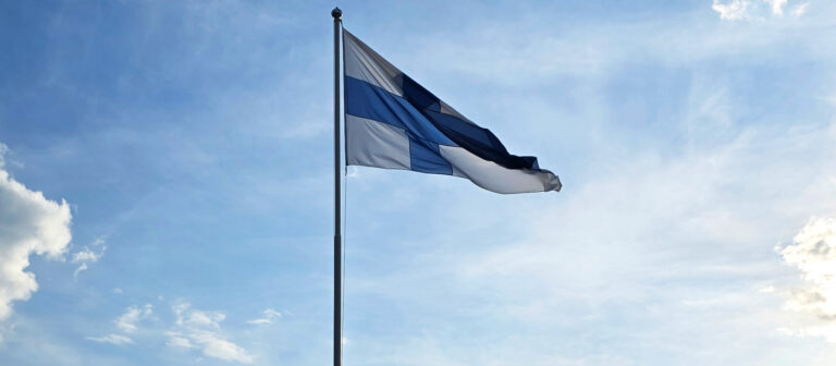 Suomen lippu liehuu, taustalla sininen taivas.