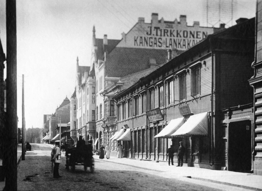 Mustavalkoisessa valokuvassa katu, jonka varrella rakennuksia. Ihmisiä ja hevoskärryt kulkee kadulla.
